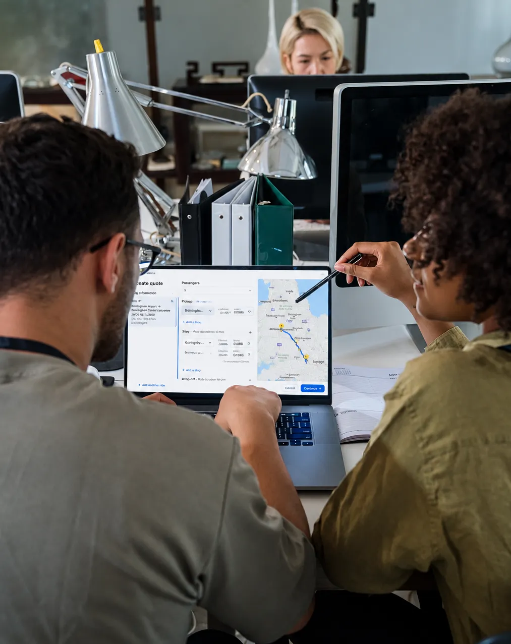 Two colleagues at a desk in a coach operator's office, seen from behind, building a quote in Mobifer on a laptop — passenger count, pickup and stops down the left, the journey drawn on a map of Britain on the right, one of them pointing at it with a pen — while a third colleague works at a monitor across the desk